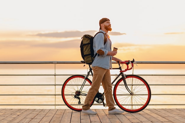 Schöner bärtiger Mann im Hipster-Stil mit Rucksack, der Jeanshemd und Mütze mit Fahrrad im Morgensonnenaufgang durch das Meer trägt, das Kaffee trinkt, gesunder aktiver Lebensstil reisender Rucksacktourist