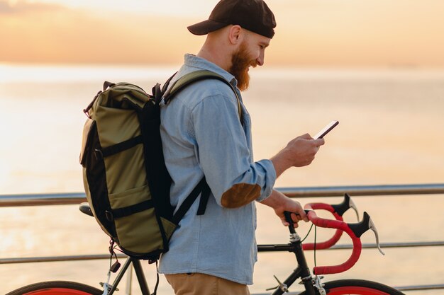 Schöner bärtiger Mann im Hipster-Stil mit Rucksack, der Jeanshemd und Mütze trägt, die mit Fahrrad im Morgensonnenaufgang durch das Meer reisen, das Kaffee trinkt, gesunder aktiver Lebensstil reisender Rucksacktourist