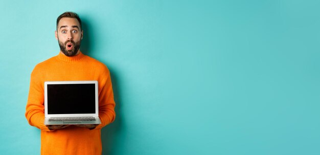 Schöner bärtiger Mann in orangefarbenem Pullover mit Laptop-Bildschirm, der Promo demonstriert, das über Licht steht