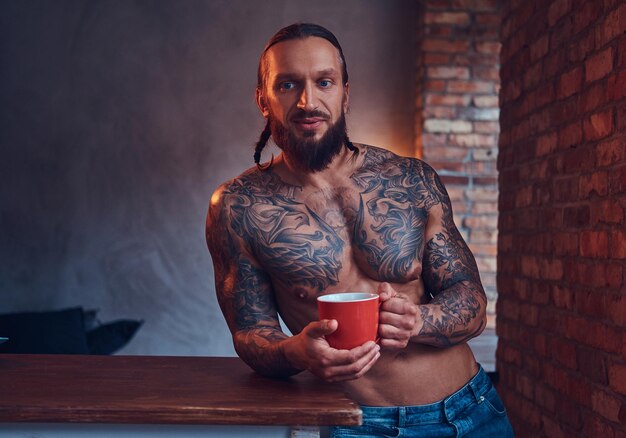 Schöner bärtiger, tätowierter Mann mit stilvollem Haarschnitt und muskulösem Körper, trinkt Kaffee und lehnt sich an einen Tisch.