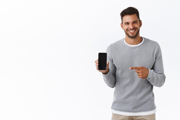 Schöner, enthusiastischer lächelnder Mann mit Borsten in grauem Pullover, der den Smartphone-Bildschirm mit Blick auf die Kamera hält und auf das mobile Display zeigt und grinst, empfiehlt die Anwendung Carsharing oder Shopping-Site