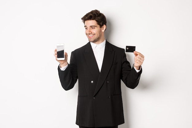 Schöner erfolgreicher Geschäftsmann, der auf den Smartphone-Bildschirm schaut und die Kreditkarte zeigt, im schwarzen Anzug vor weißem Hintergrund stehend