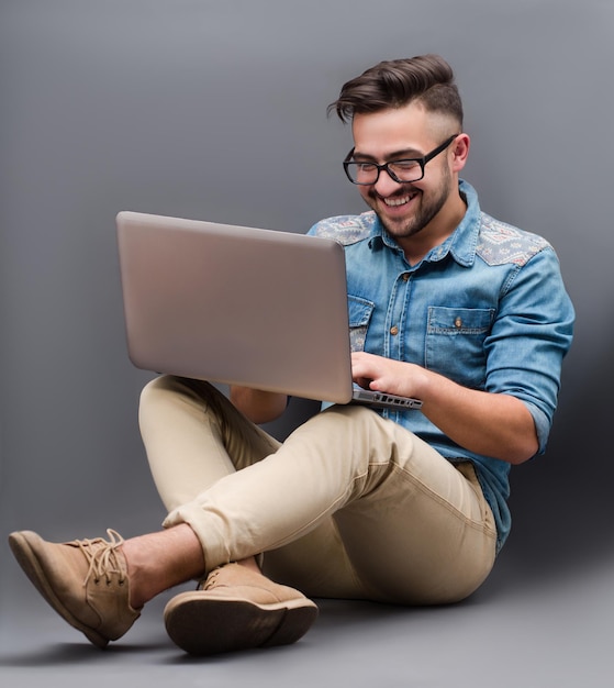 Schöner freiberuflicher Hipster-Mann, der mit seinem Laptop auf dem Boden sitzt Bärtiger Mann mit Brille, der lächelt und auf den Bildschirm schaut