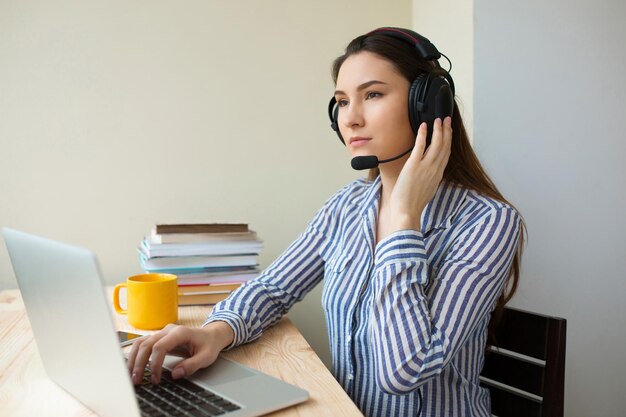 Schöner freiberuflicher Operator, der im Online-Marketing mit Headsets und einem Laptop auf einem Desktop im Büro arbeitet. Fröhliche Call-Center-Frau, die von zu Hause aus arbeitet und mit dem Kunden spricht