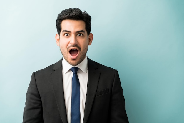 Schöner hispanischer Geschäftsmann, der mit offenem Mund vor Schock steht, während er im Studio Augenkontakt herstellt