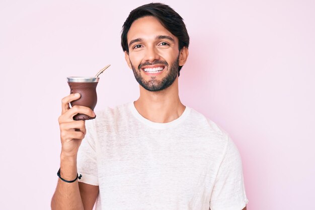 Schöner hispanischer Mann, der Mate-Infusion trinkt, sieht positiv und glücklich aus und lächelt mit einem selbstbewussten Lächeln, das Zähne zeigt