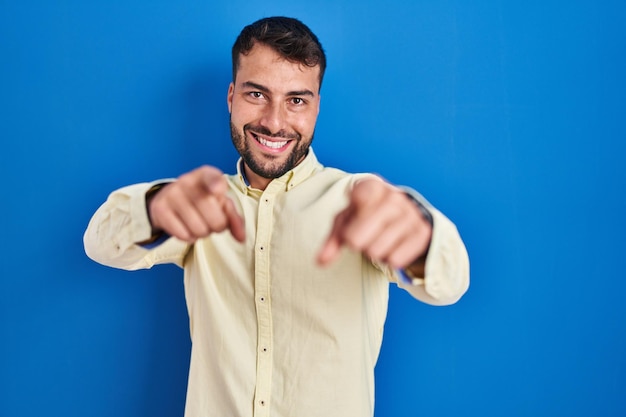 Schöner hispanischer Mann, der über blauem Hintergrund steht und mit den Fingern auf Sie und die Kamera zeigt, positiv und fröhlich lächelt