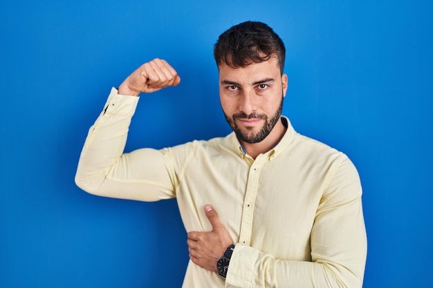 Schöner hispanischer Mann, der vor blauem Hintergrund steht, starke Person, die Armmuskeln zeigt, selbstbewusst und stolz auf Macht