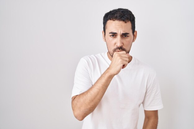 Schöner hispanischer Mann, der vor weißem Hintergrund steht und sich unwohl fühlt und hustet als Symptom für Erkältung oder Bronchitis. Gesundheitskonzept