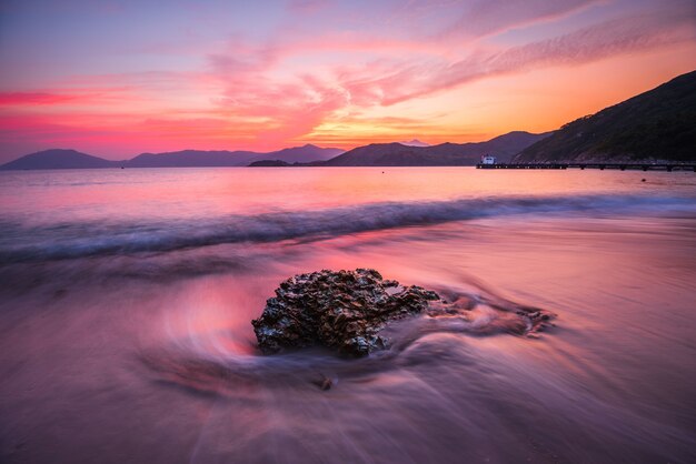 Schöner hoher Winkelschuss eines Felsens in einem welligen Meer unter einem orange und rosa Himmel bei Sonnenuntergang