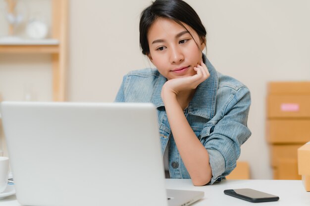 Schöner intelligenter asiatischer JungunternehmerGeschäftsfrauinhaber von KMU, das Produkt auf Lager online überprüft und zum Computer speichert, der zu Hause arbeitet.