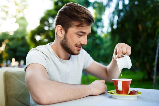Schöner junger Mann, der Sahne in Kaffee gießt, der am Tisch des Open-Air-Cafés auf verschwommenem Außenbereich sitzt