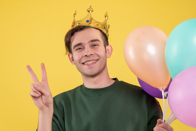 Schöner junger Mann der Vorderansicht mit der Krone, die Ballons hält, die Siegeszeichen auf gelb machen