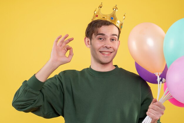 Schöner junger Mann der Vorderansicht mit Krone, die Ballons hält, die okey Zeichen auf Gelb machen