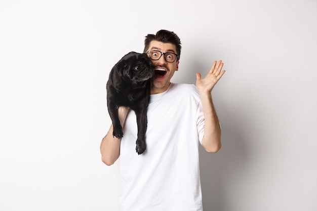 Schöner junger Mann mit Brille, der seinen schwarzen Mops hält und die Hand winkt, Typ, der Hallo sagt, während er den Hund mit einem Arm trägt und auf weißem Hintergrund steht