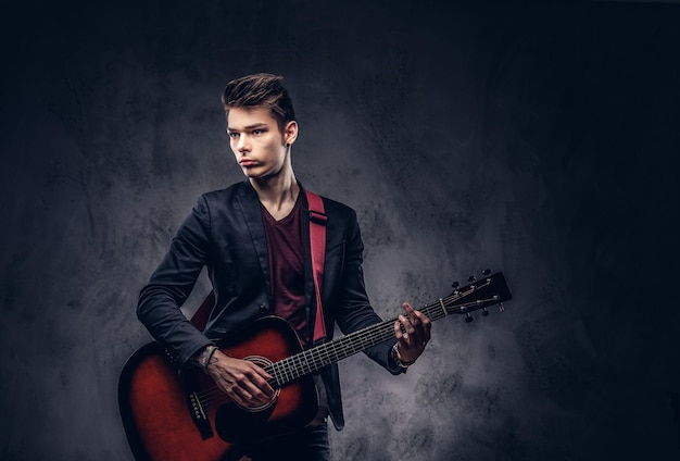 Schöner junger Musiker mit stylischem Haar in eleganter Kleidung mit einer Gitarre in den Händen, die auf dunklem Hintergrund spielt und posiert.