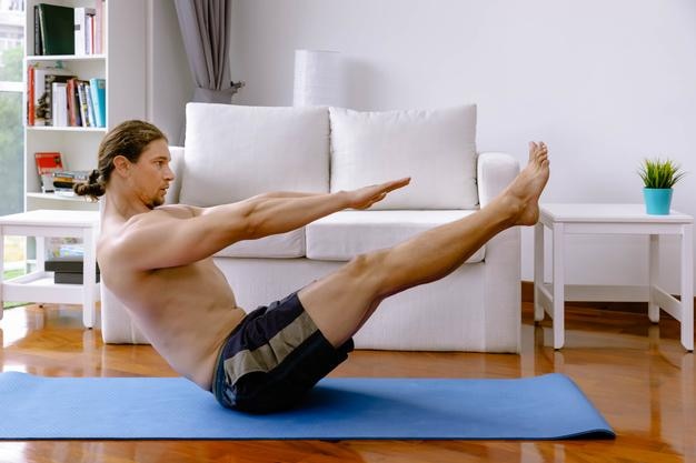 Schöner kaukasischer Mann in Sportkleidung, der Yoga praktiziert und im Wohnzimmer zu Hause eine Bootsbalance-Pose macht