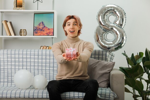 Schöner Kerl am glücklichen Frauentag, der auf dem Sofa im Wohnzimmer ein Geschenk vor der Kamera hält