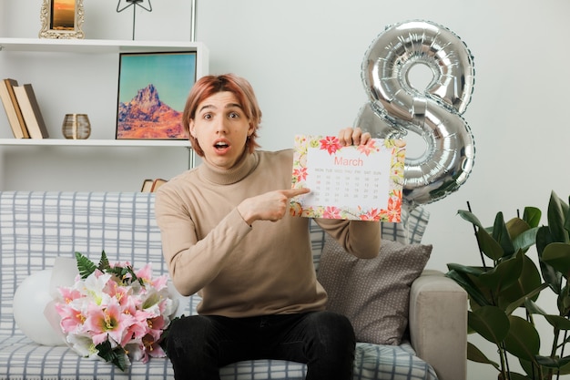 Schöner Kerl am glücklichen Frauentag, der auf dem Sofa im Wohnzimmer sitzt und auf den Kalender punktet