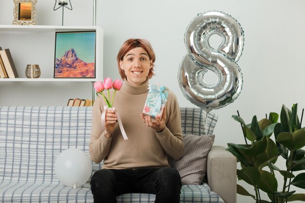 Schöner Kerl am glücklichen Frauentag, der Blumen mit Geschenk auf dem Sofa im Wohnzimmer hält