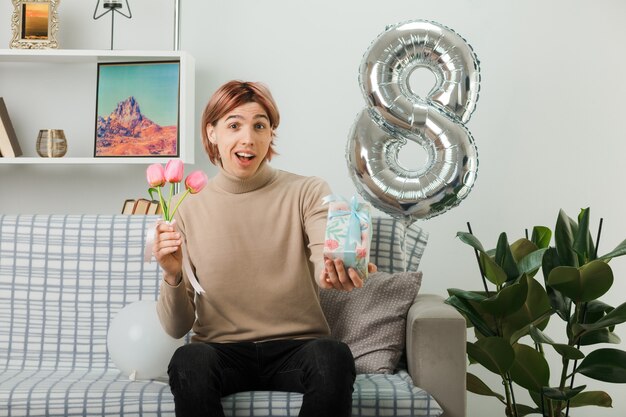 Schöner Kerl am glücklichen Frauentag, der Blumen mit Geschenk auf dem Sofa im Wohnzimmer hält