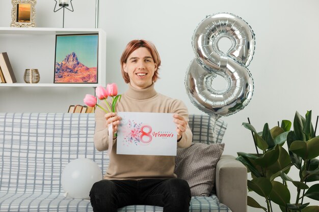 Schöner Kerl am glücklichen Frauentag, der Blumen mit Postkarte in die Kamera hält, die auf dem Sofa im Wohnzimmer sitzt
