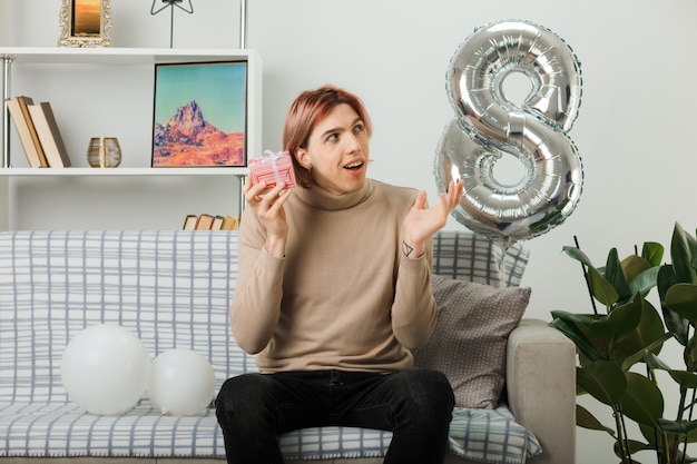 Schöner Kerl am glücklichen Frauentag, der das Geschenk auf dem Sofa im Wohnzimmer hält