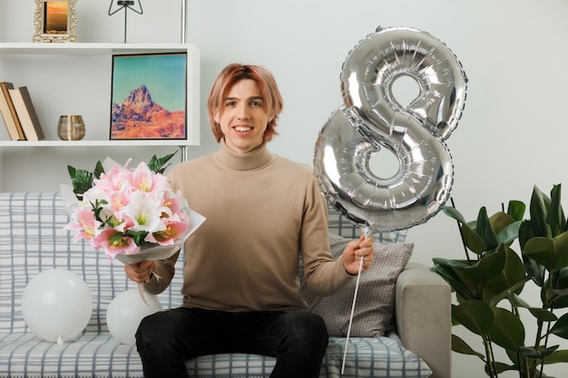 Schöner Kerl am glücklichen Frauentag, der den Ballon Nummer acht und den Blumenstrauß hält, der auf dem Sofa im Wohnzimmer sitzt