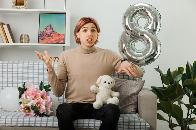 Schöner Kerl am glücklichen Frauentag mit Teddybär, der auf dem Sofa im Wohnzimmer sitzt
