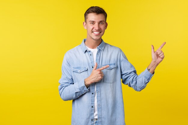 Schöner lächelnder glücklicher Mann mit weißen Zähnen, der mit den Fingern nach rechts zeigt, Kunden einlädt, Werbung zu sehen, neue Produkte zu demonstrieren, spezielle Rabattangebote, stehender gelber Hintergrund
