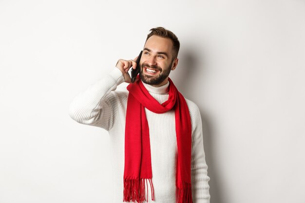 Schöner lächelnder Mann, der am Telefon spricht, zufrieden aussieht, im Winterschal und Pullover steht, weißer Hintergrund