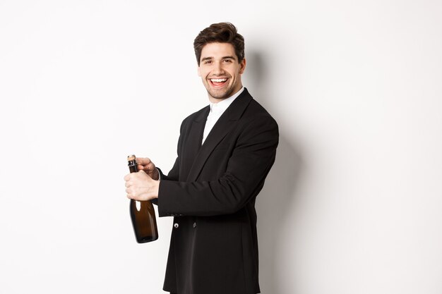 Schöner lächelnder Mann im trendigen Anzug öffnet eine Flasche Champagner, feiert Feiertage und steht auf weißem Hintergrund