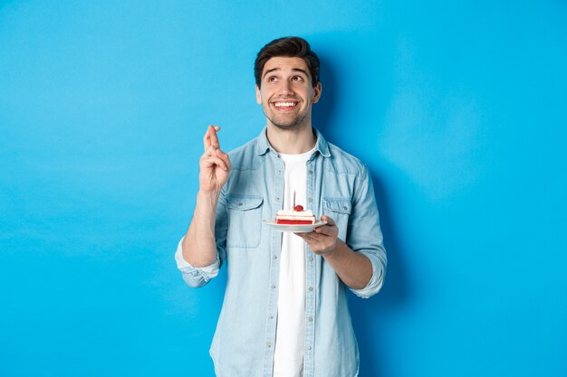 Schöner Mann, der einen Wunsch macht, Geburtstag feiert, B-Day-Kuchen hält und die Finger kreuzt und auf blauem Hintergrund steht