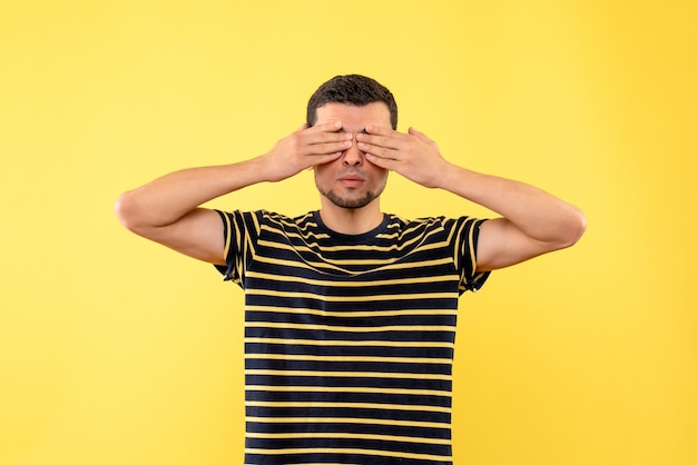 Schöner mann der vorderansicht im schwarz-weiß gestreiften t-shirt, das augen mit händen auf gelbem lokalisiertem hintergrund bedeckt