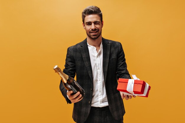 Schöner Mann in stilvollem kariertem Anzug und trendigem weißem Hemd zwinkert lächelnd und hält Champagnerflasche und Geschenkbox auf orangefarbenem Hintergrund