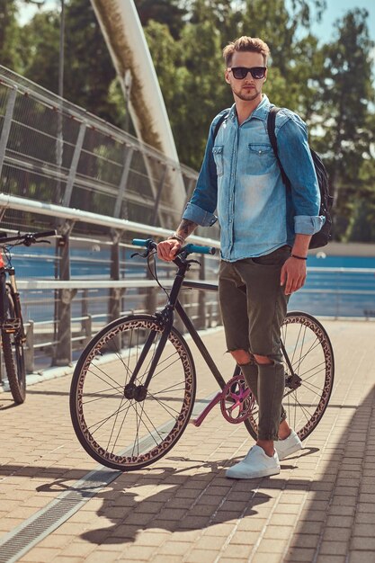 Schöner modischer Hipster mit stylischem Haarschnitt in Sonnenbrille, der mit dem Fahrrad im Freien spazieren geht.