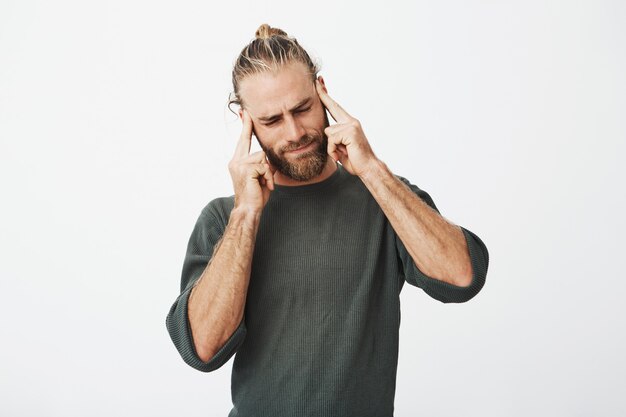 Schöner nordischer Mann mit trendigem Haarschnitt und Bart, der müde ist und Schläfen mit den Fingern massiert, die versuchen, sich zu konzentrieren