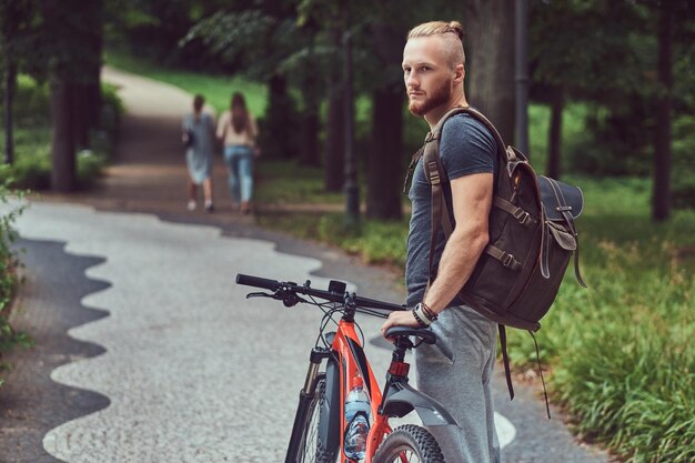 Schöner rothaariger Mann mit stylischem Haarschnitt und Bart in Freizeitkleidung spaziert mit Fahrrad und Rucksack im Park.