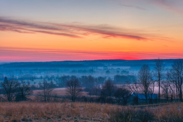 Schöner Schuss von erstaunlichen roten und orange Wolken über großen nebligen Feldern und Wald im Morgengrauen