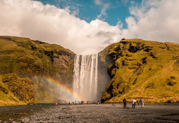 Schöner Schuss von Leuten, die nahe dem Wasserfall mit einem Regenbogen auf der Seite in Skogafoss Island stehen