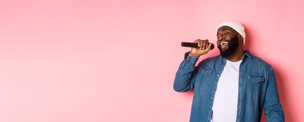Schöner schwarzer Mann in Mütze und Jeanshemd, der Karaoke singt und das Mikrofon hält, das über rosa steht b