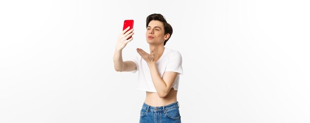 Schöner schwuler Mann im bauchfreien Oberteil, der einen Luftkuss an der Telefonkamera sendet und ein Selfie oder einen Video-Chat auf Smartp macht