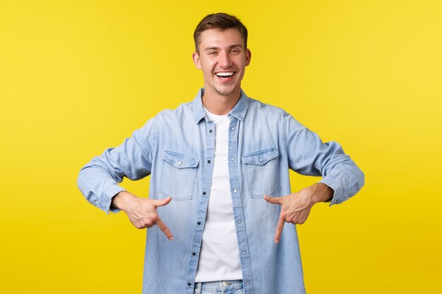 Schöner selbstbewusster lächelnder Mann in lässigem Outfit, der mit den Fingern nach unten zeigt, um Werbung zu zeigen, Klickbanner empfehlen, Sonderangebot im Geschäft, gelber Hintergrund glücklich stehen.