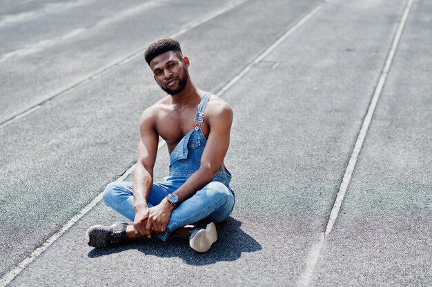 Schöner sexy afrikanisch-amerikanischer Mann mit nacktem Oberkörper in Jeans-Overalls, der auf der Rennbahn des Stadions sitzt Modisches Porträt eines schwarzen Mannes