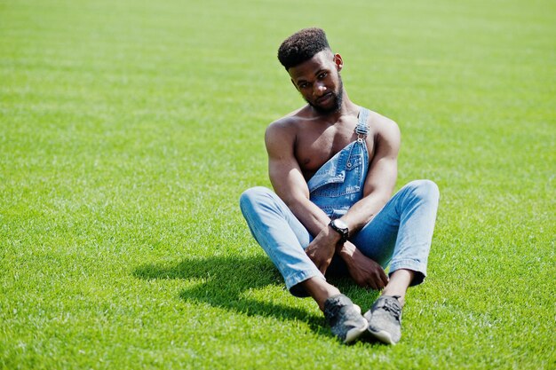 Schöner sexy afrikanisch-amerikanischer Mann mit nacktem Oberkörper in Jeans-Overalls, der auf grünem Gras des Stadion-Fußballplatzes sitzt Modisches Porträt eines schwarzen Mannes