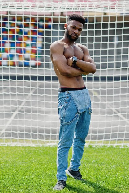 Schöner sexy afroamerikanischer muskulöser Mann mit nacktem Oberkörper in Jeans-Overalls, der auf grünem Gras gegen Fußballtore des Stadionfeldes posiert Modisches Porträt eines schwarzen Mannes