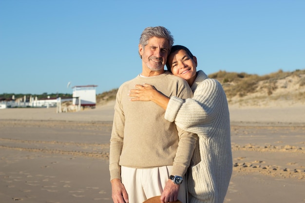 Schönes älteres Paar, das am Strand steht und für die Kamera posiert. Schöner grauhaariger Ehemann und Ehefrau, die einen warmen Herbstabend an der Küste genießen, lächeln und sich umarmen. Liebe, Ruhestandskonzept