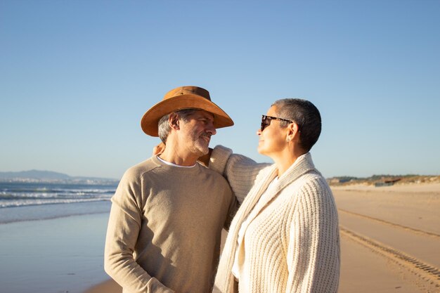 Schönes älteres Paar, das an der Küste steht, sich umarmt und ansieht. Mann mit Cowboyhut und kurzhaarige Frau mit Sonnenbrille genießen gemeinsam Urlaub am Meer. Freizeit, Ruhestandskonzept