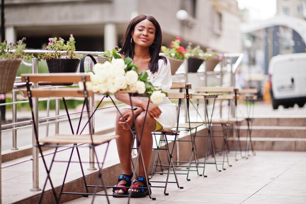 Schönes afrikanisch-amerikanisches Mädchen, das Blumenstrauß aus weißen Rosen bei der Datierung in der Stadt hält Schwarze Geschäftsfrau mit Blumenstrauß, die am Tisch im Café im Freien sitzt