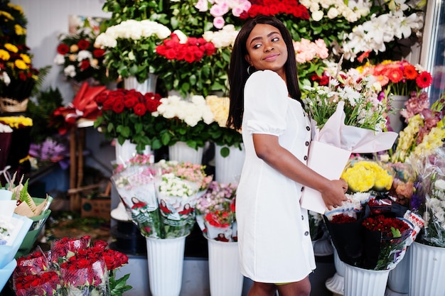 Schönes afrikanisch-amerikanisches Mädchen in zartem weißem Kleid mit Blumenstrauß in den Händen, die vor floralem Hintergrund im Blumenladen stehenSchwarze Floristin